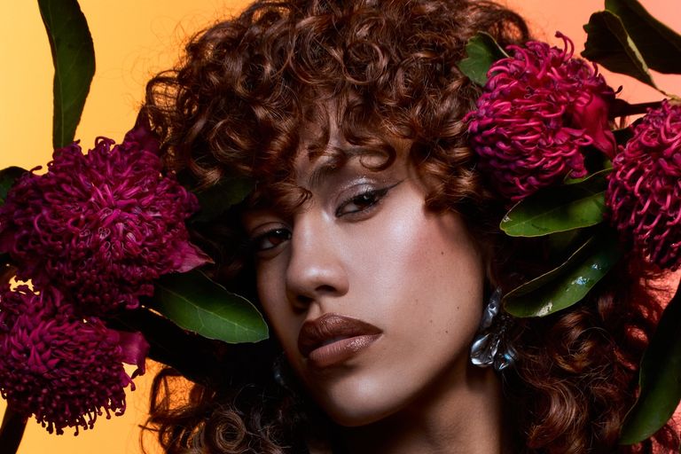 Close-up portrait of musician Becca Hatch with curly hair framed by bright pink native Australian flowers against a warm yellow background.